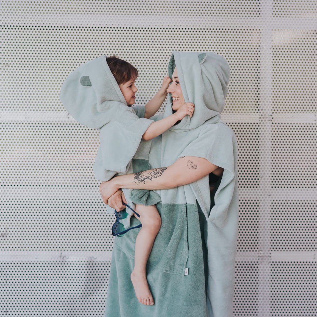 1. Adult and child wearing matching green Surfbear bath ponchos with hoods and ears, smiling at each other, in front of a metal background