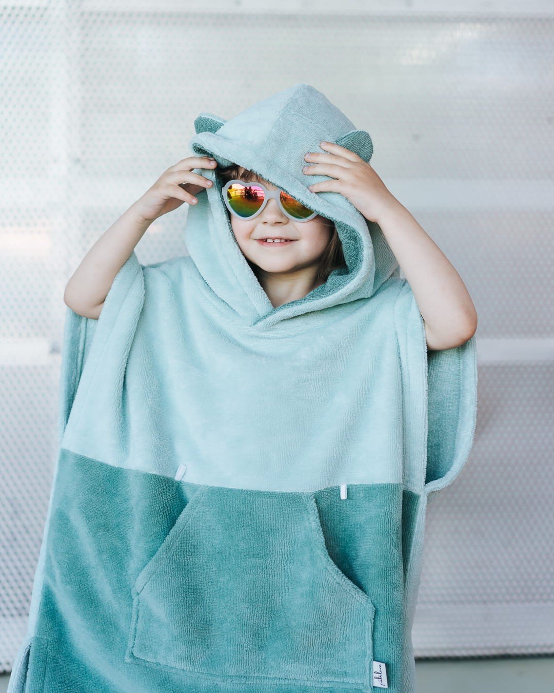 1. Child wearing green Surfbear kids bath poncho with hood and ears, smiling and holding hood, in front of a metal background