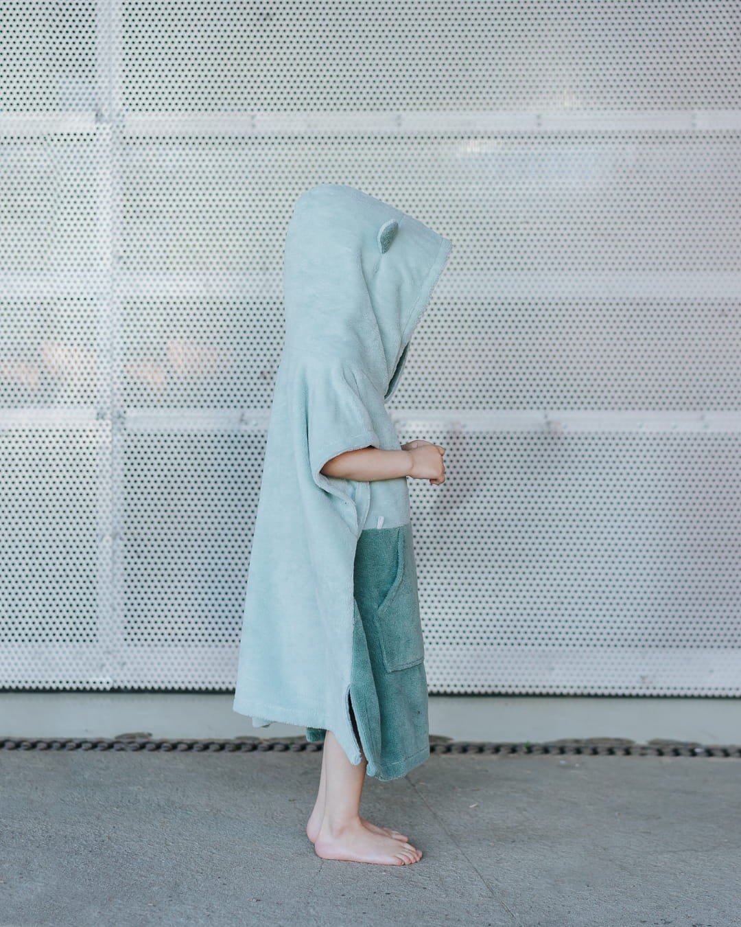 1. Side view of child wearing green Surfbear kids bath poncho with hood and ears, standing in front of a metal background
