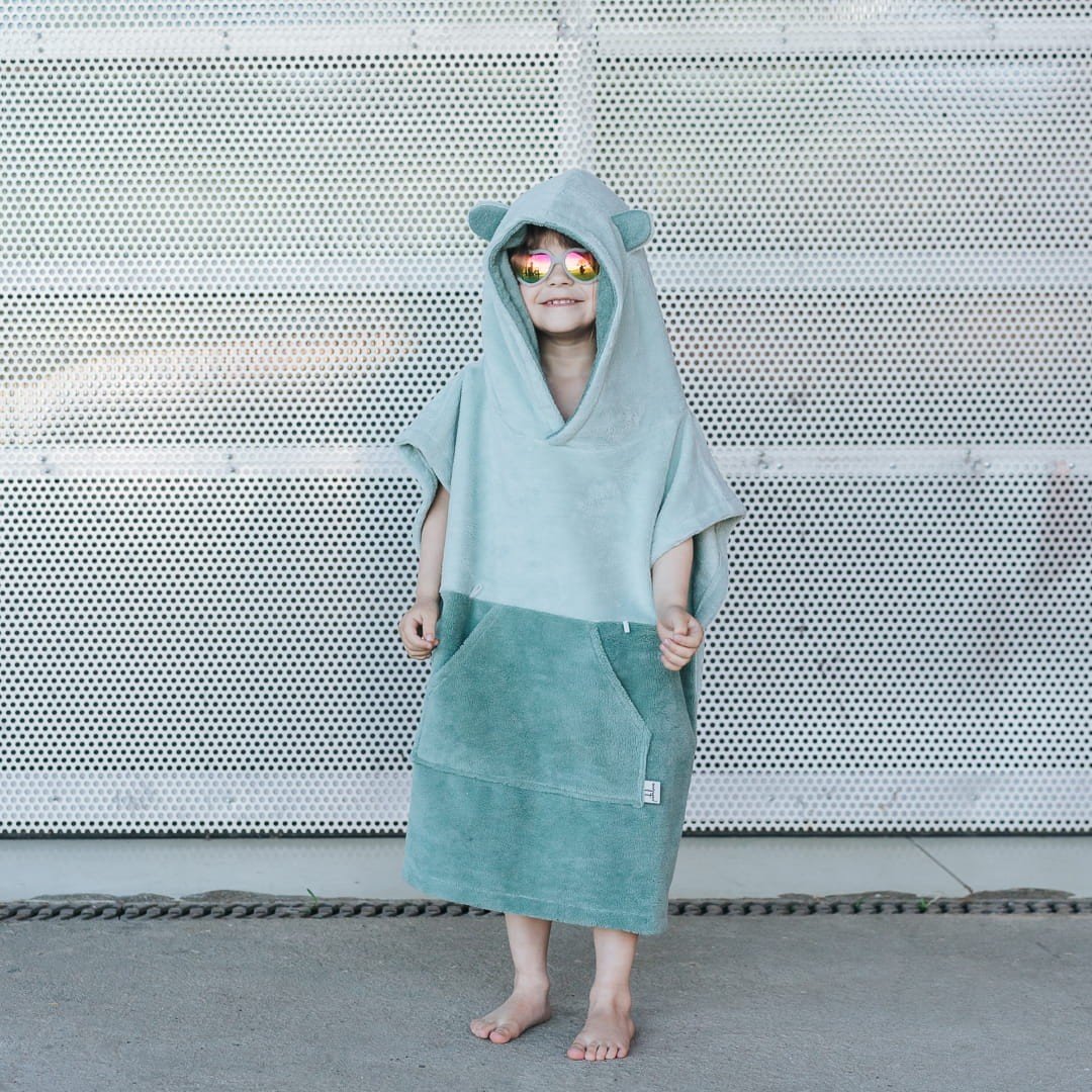 1. Child standing in green Surfbear kids bath poncho with hood and ears, smiling, in front of a metal background