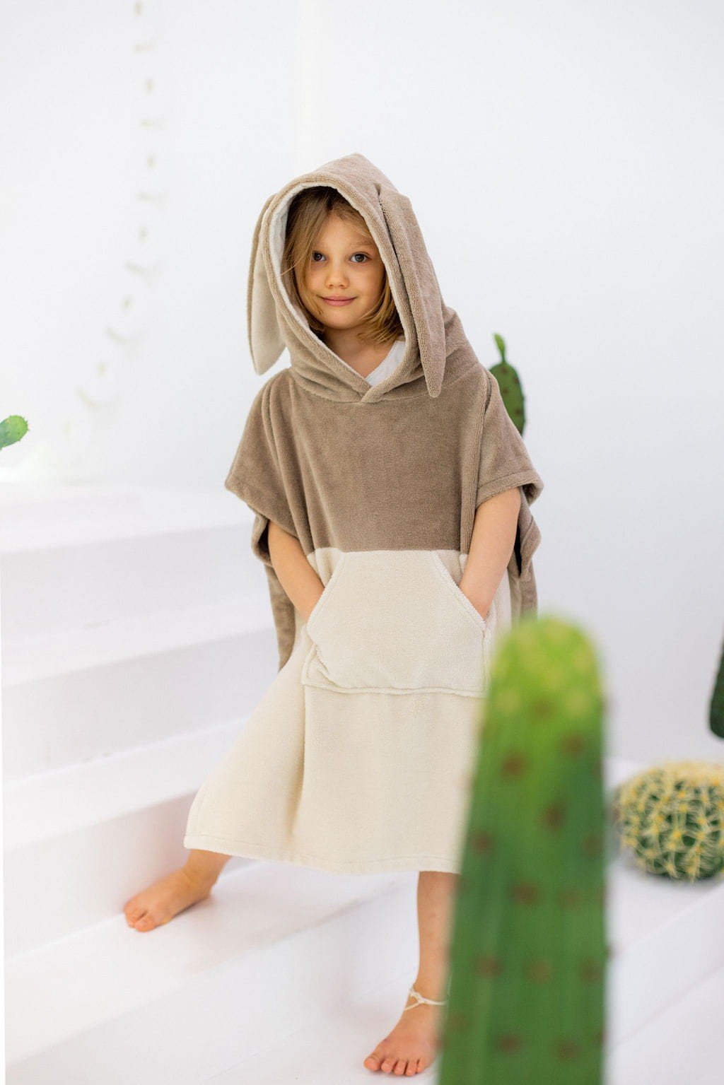 1. Child wearing Patulove Surfbunny kids bath poncho in latte color with hood and kangaroo pocket, standing on white steps with cacti in background