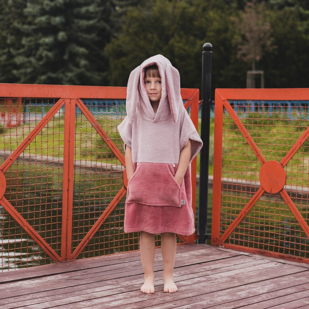 1. Child in pink Surfbunny bath poncho with bunny ears standing on wooden deck by water