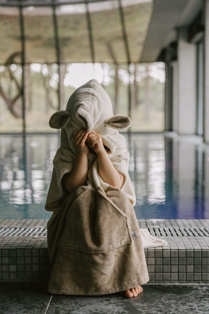 4. Child sitting by indoor pool wearing beige Surfsheep kids bath poncho with hood and ears covering face