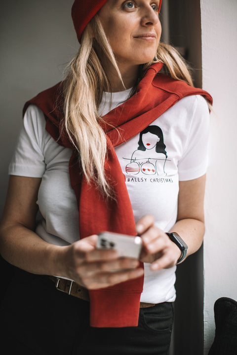 1. Woman wearing white 'Ballsy Christmas' T-shirt with red sweater draped over shoulders, holding phone, standing by window