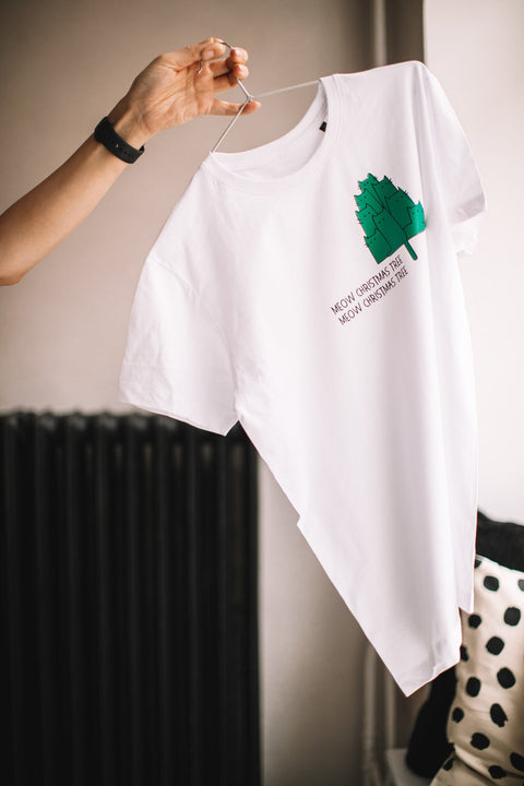 2. White T-shirt with green Christmas tree graphic and 'Meow Christmas Tree' text held up by hand in a minimalist room