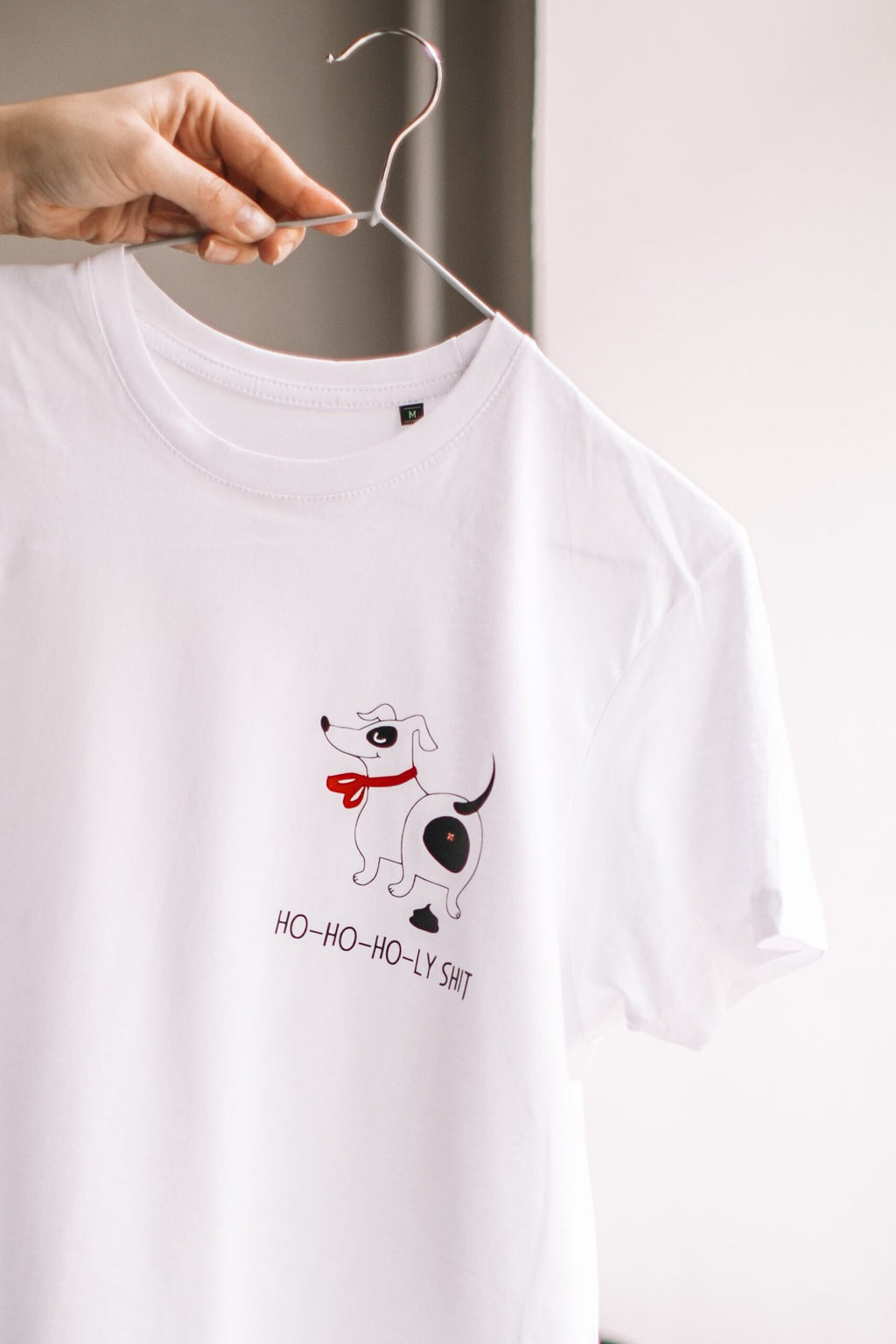 2. White T-shirt with dog graphic and 'Ho-ho-ho-ly sh*t' text held on hanger, showcasing design