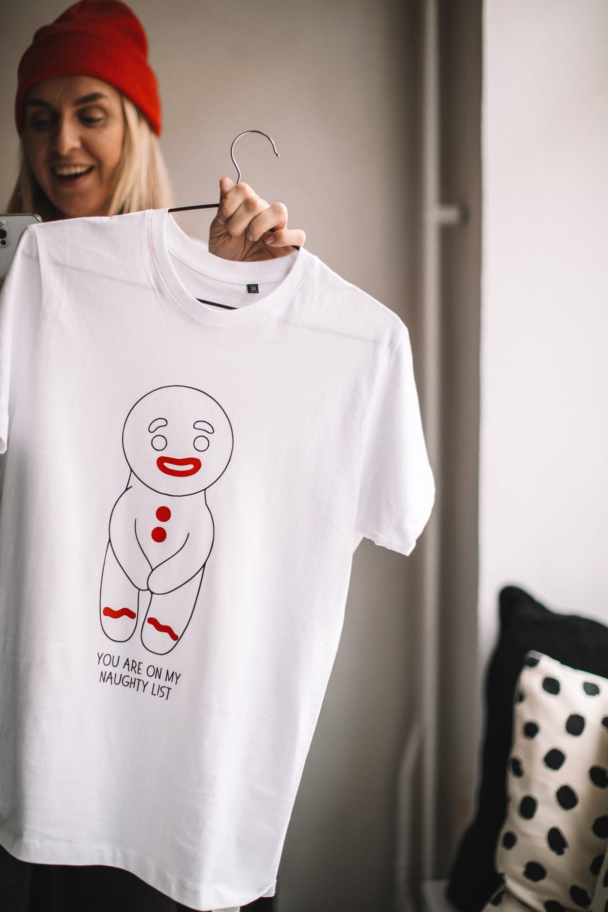 2. Woman in red beanie displaying white T-shirt featuring gingerbread man design and 'You are on my naughty list' text, indoors