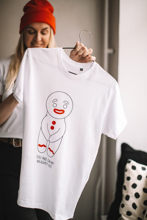 1. Woman in red beanie holding white T-shirt with gingerbread man graphic and 'You are on my naughty list' text, standing in cozy room