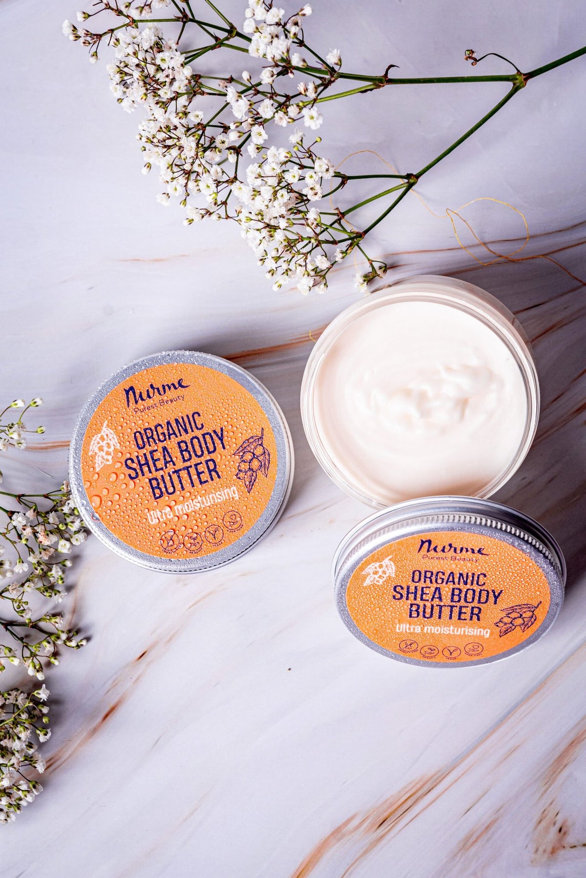 1. Open and closed Nurme shea body butter containers on marble surface with white flowers