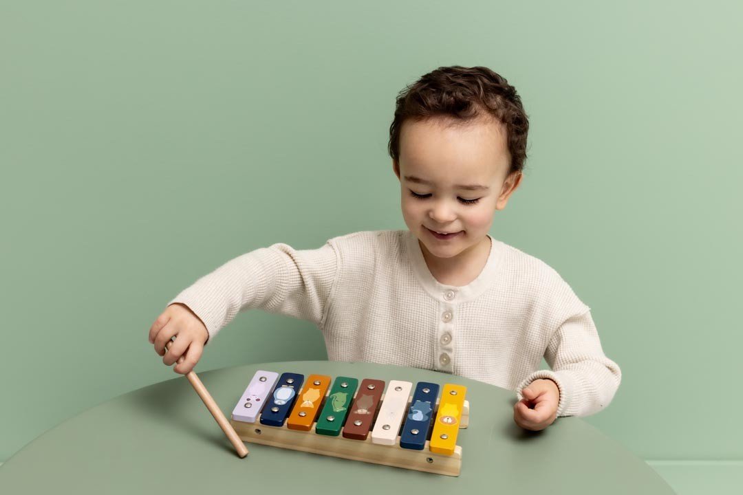 1. Child playing with Trixie Baby wooden xylophone on green table, wearing cream sweater, smiling