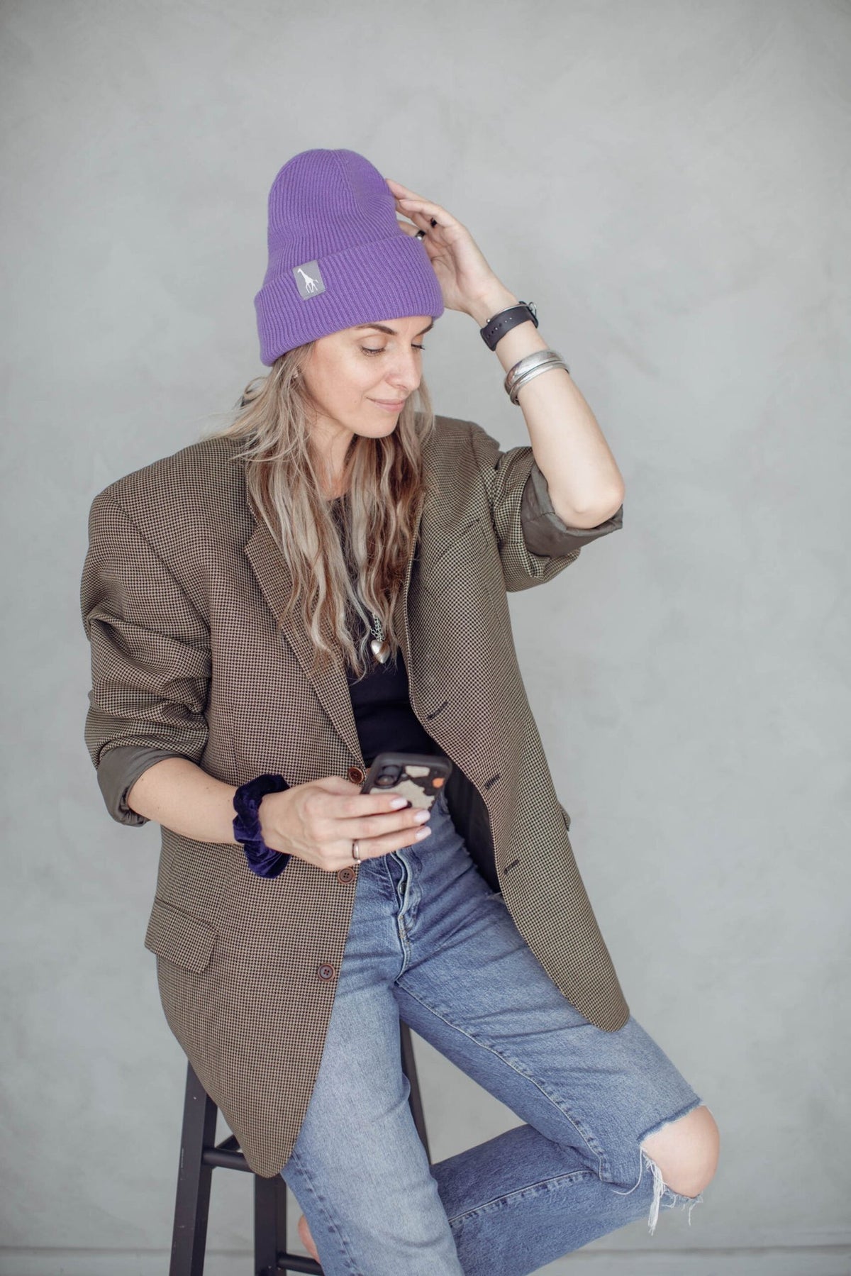1. Woman wearing purple beanie with giraffe label, sitting on stool in casual outfit, holding phone