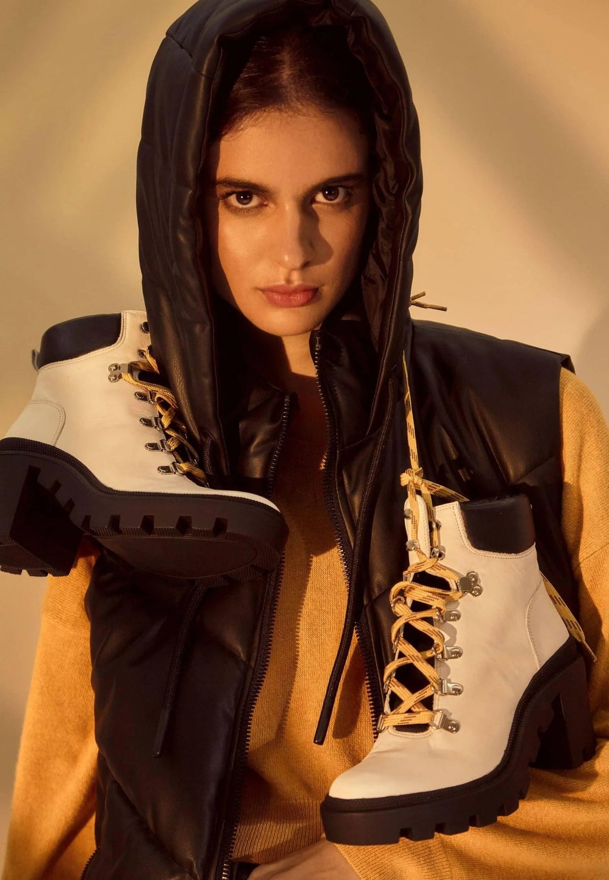 1. Woman holding white combat boots with 9 cm block heel and yellow laces, wearing black vest and hood in warm-toned setting