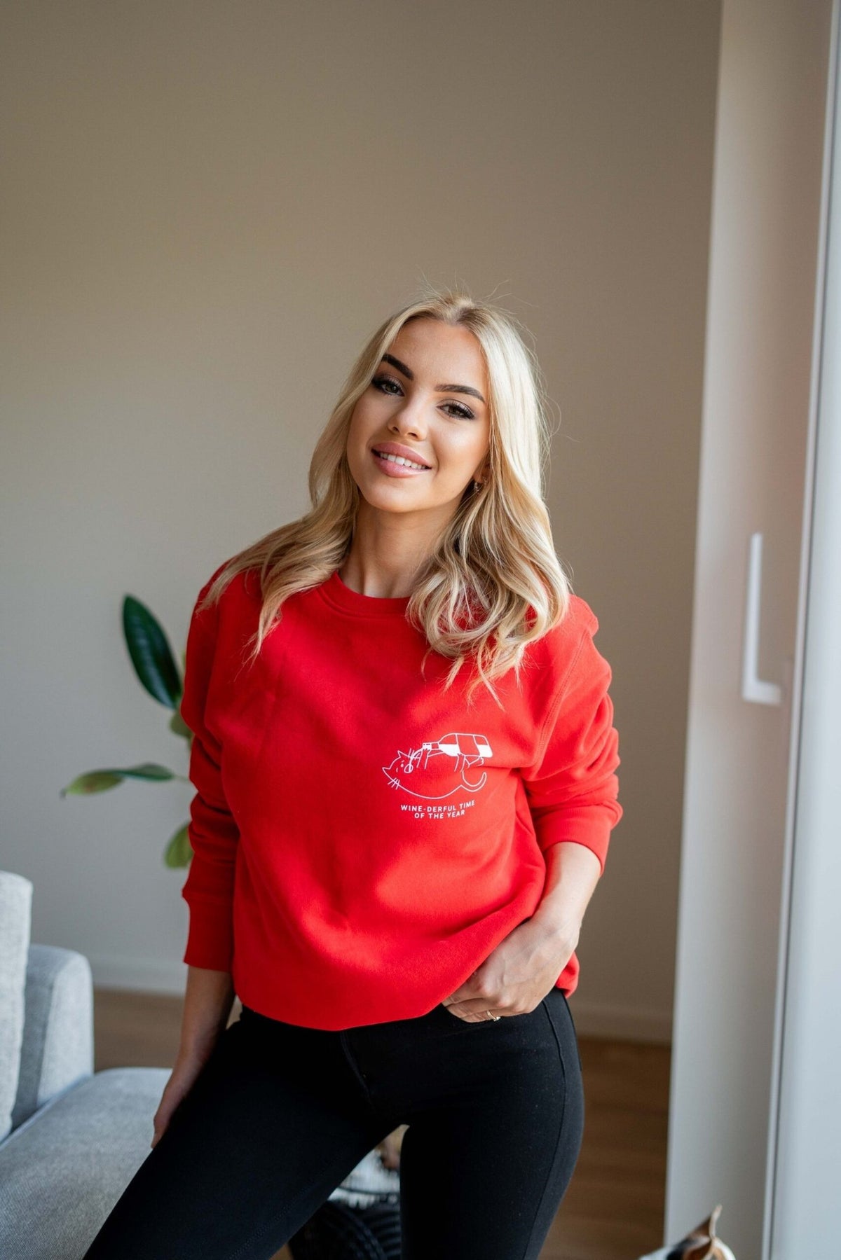 2. Woman smiling in red oversized sweatshirt with white graphic and text 'Wine-derful Time of the Year' standing in a bright room