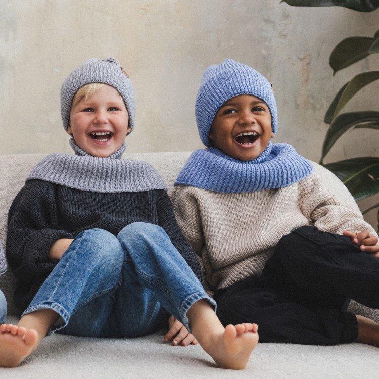 3. Two children sitting on sofa, one wearing grey and the other sky blue merino wool hat and scarf, laughing