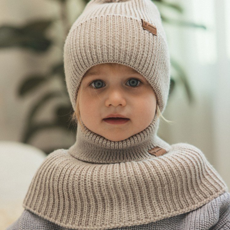 2. Child wearing Patulove oat merino wool tube scarf and hat, sitting indoors with a neutral expression