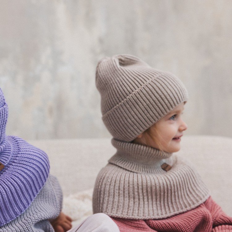 4. Two children sitting indoors, one wearing oat-colored merino wool tube scarf and hat, the other in purple