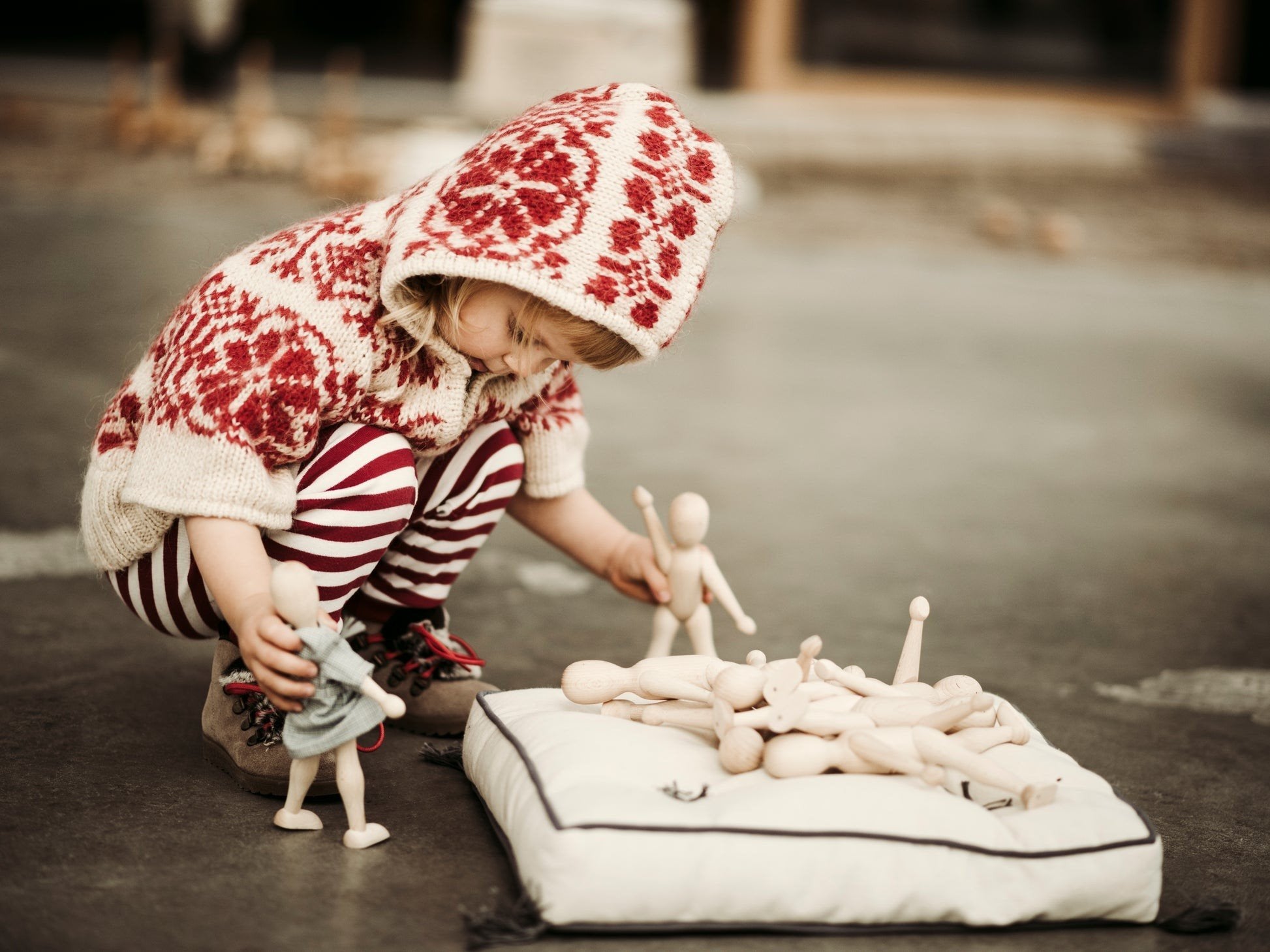 9. Child in red patterned sweater arranging Wooden Story beech dolls on cushion outdoors