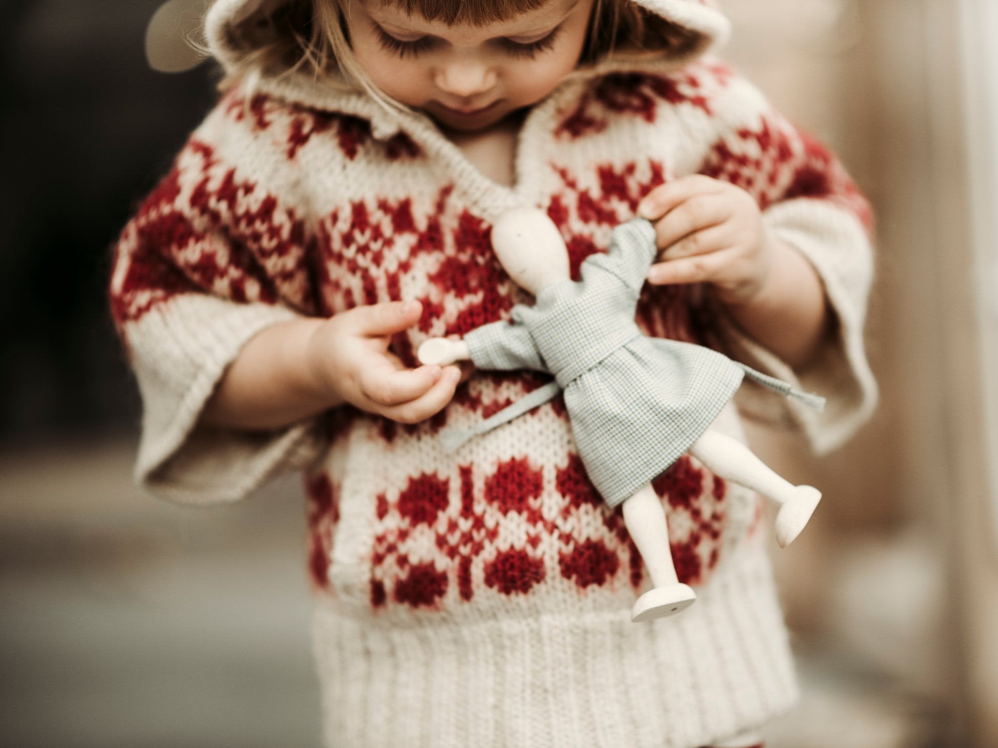 1. Child in red patterned sweater holding Wooden Story beech doll with grey dress outdoors