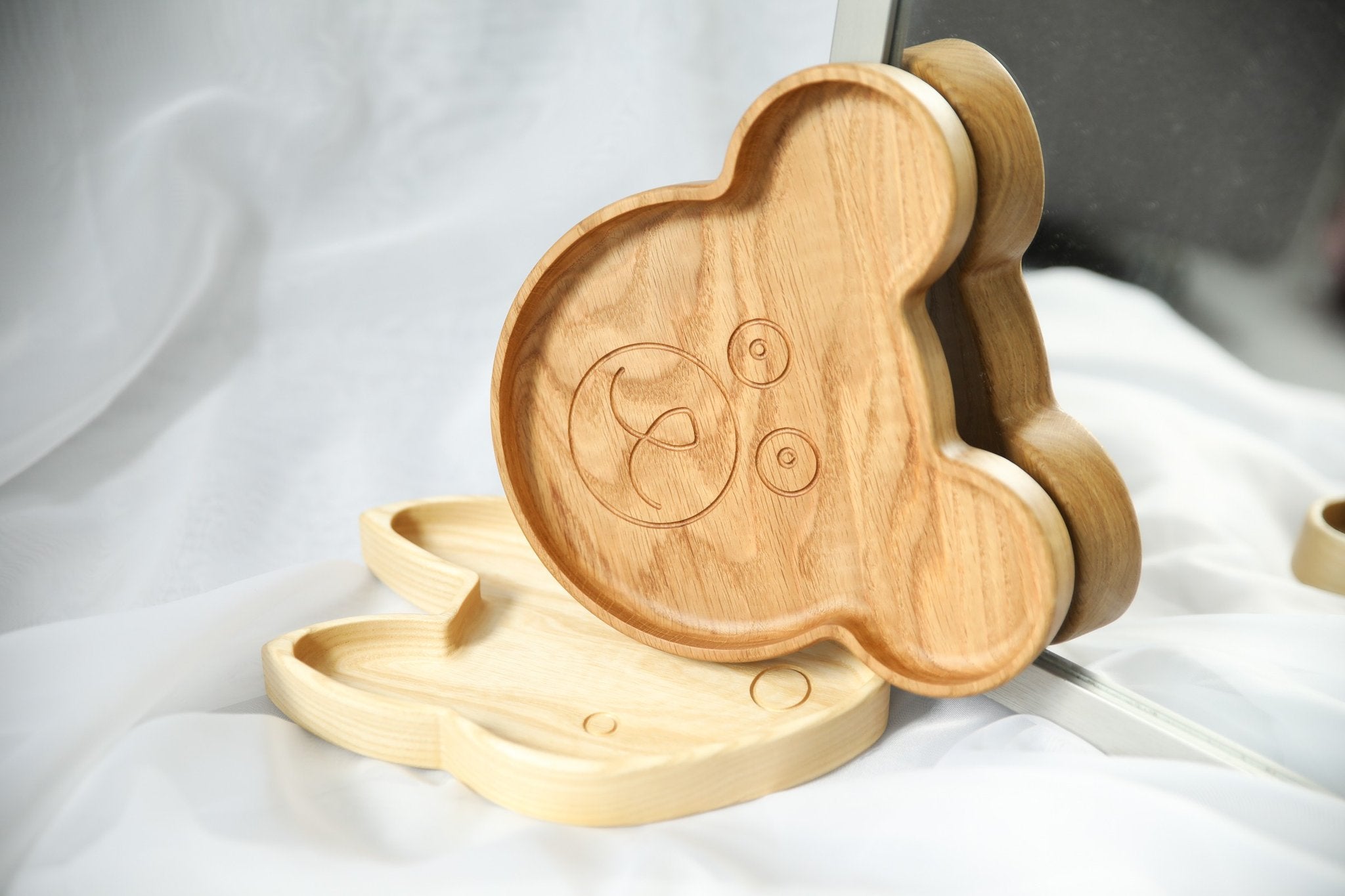2. Two wooden bear-shaped plates with engraved face design, displayed on white fabric