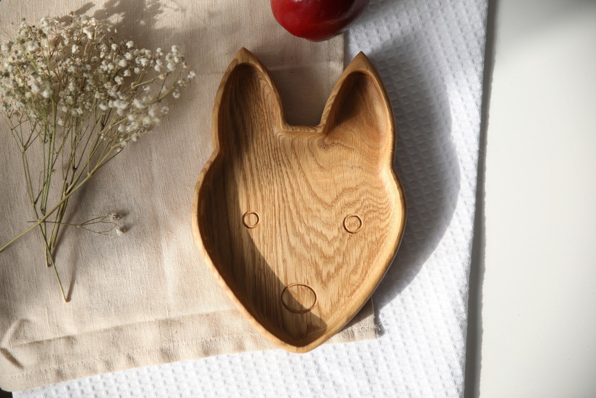 4. Front view of wooden fox-shaped plate on beige cloth with red apple and white flowers, highlighting engraved face details